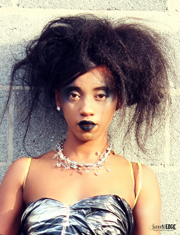 Closeup of model standing against cinder block wall wearing trash bag dress and saftey pin necklace.